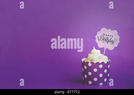 Vainilla cupcake con pequeños corazones decorativos y feliz cumpleaños a firmar, en violeta sobre papel de hornear de Copa, contra el fondo violeta Foto de stock