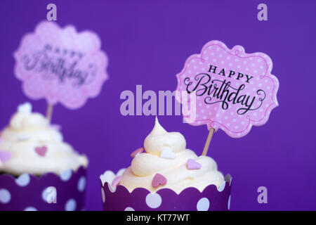 Cupcakes de vainilla con pequeños corazones decorativos y feliz cumpleaños firmar, contra el fondo violeta; Partido antecedentes Foto de stock