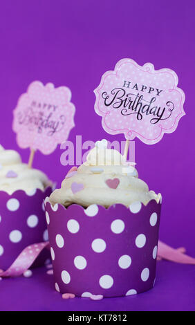Cupcakes de vainilla con pequeños corazones decorativos y feliz cumpleaños firmar, contra el fondo violeta; Partido antecedentes Foto de stock