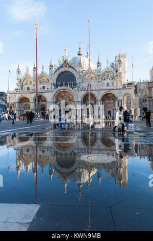 La Basílica de San Marcos se refleja en acqua alta en Piazza San Marco con turistas, wheeling maletas Venecia, Véneto, Italia