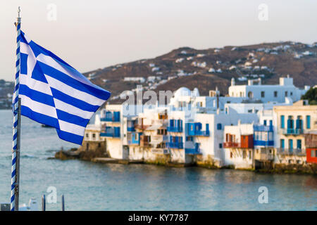 Ondear la bandera griega en el verano con vistas del atardecer reflejándose en Little Venice, Mykonos, Grecia, verano de 2017. Foto de stock