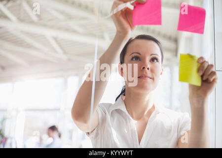Hermosa joven empresaria poniendo notas adhesivas en pared de vidrio en la oficina Foto de stock