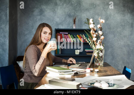 Feliz sonriente joven diseñador hermosa mujer sentada en la mesa con una taza de café, posando, mirando a la cámara. Utilización Portátil modelo alegre