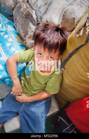Un niño filipino preescolares con grandes ojos asiáticos, cautivador y una ligera sonrisa se asienta entre bolsas de carga en un ferry en Filipinas. Foto de stock