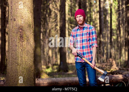 Logger barbudo en una camisa a cuadros con un hacha de pie cerca de un árbol en el bosque Foto de stock