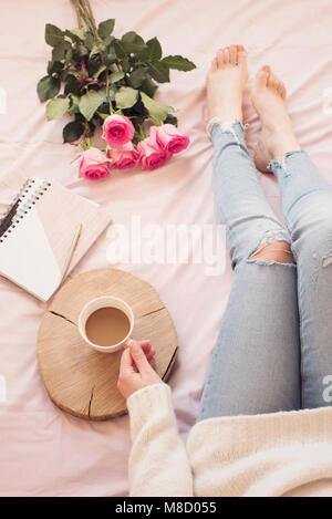 Joven mujer caucásica en la cama temprano en la mañana, vistiendo pantalones vaqueros angustiado y un suéter blanco y tiene una taza de café. Las rosas y los portátiles. Foto de stock