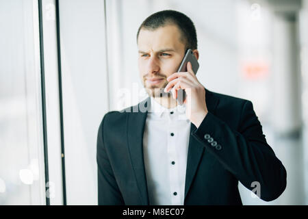 Joven empresario hablando en teléfono móvil mirando fuera de la oficina. Empresario de pie junto a la ventana de highrise edificio de oficinas. Foto de stock