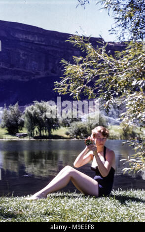 Mujer joven con traje de baño de una pieza sentada en la orilla de un lago mirando a sí misma en un espejo de bolsillo, en EE.UU. En 1950