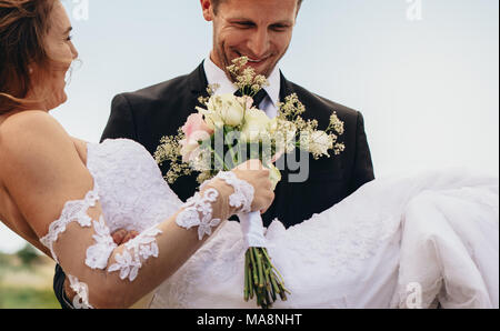 Feliz novio llevar beautiful bride con flores en la mano. Pareja de recién casados con bouquet afuera, en el día de su boda.