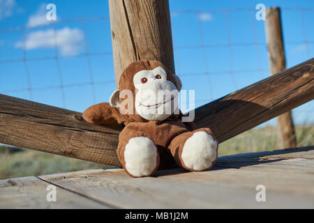 Primer plano de un precioso color marrón y blanco mono de juguete sonriendo felizmente sentado sobre un piso de madera y recostada cómodamente en una baranda de madera Foto de stock