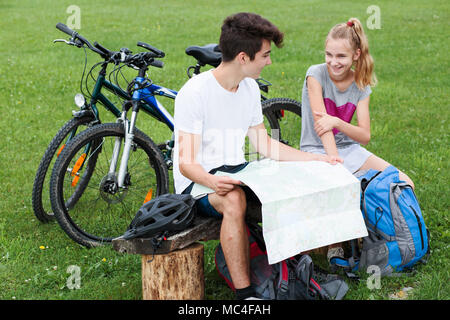 Sonriente joven chico y chica tener un descanso sentado en un banco durante el viaje en bicicleta y planificar una mayor parte del viaje con el mapa equipado con cascos backpac Foto de stock
