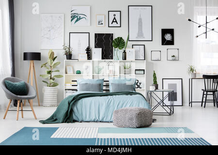 Moqueta azul y pouf cerca de la cama en el dormitorio espacioso interior con sillón gris y carteles Foto de stock
