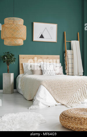 Pouf y piel blanca en el suelo cerca de la cama con manta tejida en verde esmeralda del interior del dormitorio con la planta y cartel Foto de stock