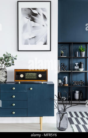 Close-up de una pintura, armario azul, radio retro y vaso de vidrio oscuro con ramificaciones en el salón interior y una estantería metálica en el fondo Foto de stock
