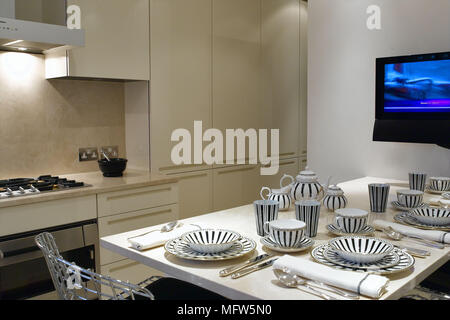 Una cocina moderna con tonos beige cocina frente a una mesa de cocina con  lugar la configuración y una gran pantalla plana de televisión empotrada en  la pared Fotografía de stock 