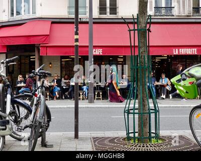 Mujer de pañuelo y largo, fluyendo ropa caminando delante de gente sentada fuera una cafetería, París, Francia.
