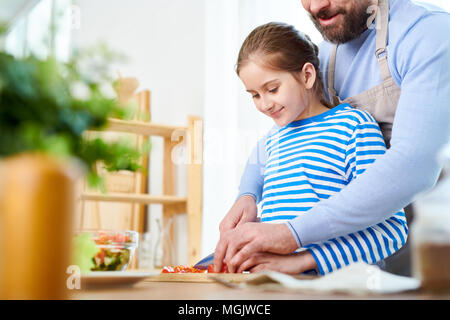 Momentos encantadores de la paternidad: sonriente niña cortando tomates para preparar ensalada de verduras mientras su papá barbudo ayudarla, interior o Foto de stock