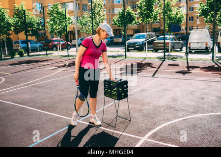 Mujer, jugador de tenis colocando pelotas de tenis en una canasta en la pista de tenis. Foto de stock