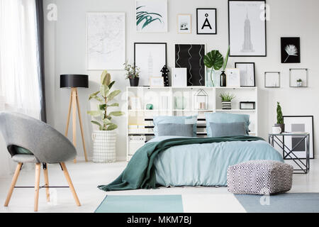 Sillón gris y telas pouf cerca de la cama con una manta azul verde en el interior del dormitorio con galería Foto de stock