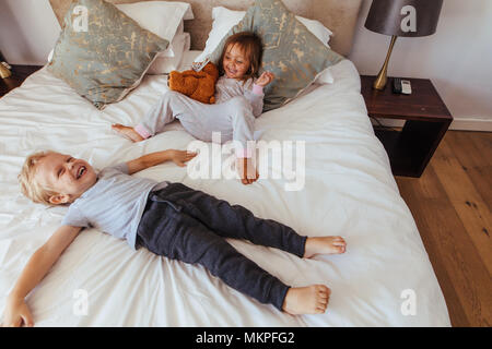 Niñito acostado en la cama con su hermana sentados sosteniendo un oso de peluche sonriendo. Poco alegres niños jugando en el dormitorio.