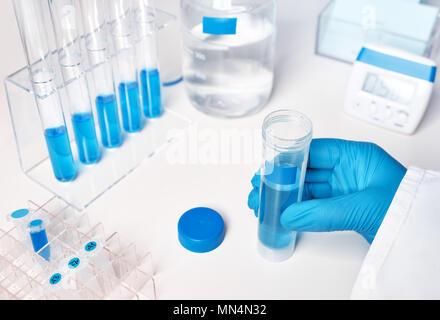 Antecedentes científicos, en azul y blanco. Muestra de líquido en el lado femenino enguantada, azul muestras líquidas en tubos de vidrio y plástico Foto de stock