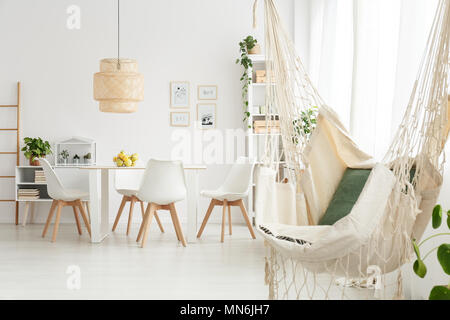 Hamaca con almohadas y posters en la pared en el comedor interior con lámpara de mimbre sobre la mesa con sillas blancas Foto de stock