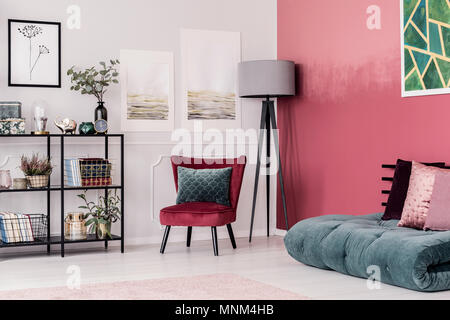 Verde esmeralda y rosa con futón almohadas en el piso contra la pared roja, ombre en el elegante salón interior con lámpara y silla de terciopelo gris Foto de stock