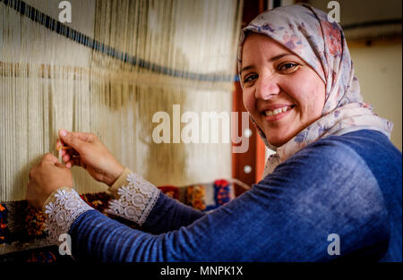 Una mujer con la vestimenta tradicional se sienta tejer alfombras en Tazenakht, sur de Marruecos, África Foto de stock