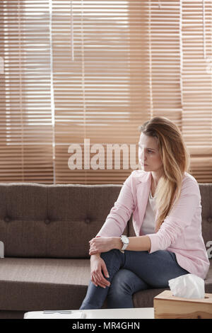 Mujer joven con depresión sentada sola en un sofá con una caja de pañuelos en frente de ella durante una sesión de terapia