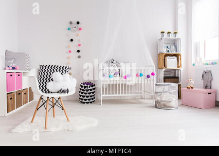Dormitorio blanco diseñado para bebé niña. Por la pared cuna con dosel, estantería y juguetes. En el oriente blanco sillón con almohadas