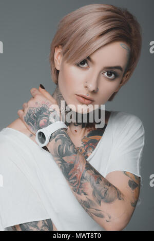 Retrato de hermosa chica tatuada en camiseta blanca mirando a la cámara aislada sobre gris Foto de stock