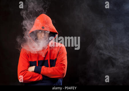Un joven con una capucha naranja de pie en posición cerrada con cabeza baja fuma un vape y exhala un humo blanco sobre un fondo negro aislado Foto de stock