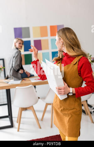Editor de la revista atractiva con carpeta apuntando a la colega y la paleta de colores en la pared detrás de Foto de stock