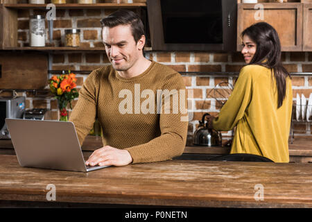 Sonriente joven con ordenador portátil mientras novia cocinando detrás en la cocina