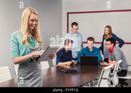 Una joven y bella mujer de negocios milenario trabajando en su tableta en una sala de conferencias con su co-trabajadores; en Sherwood Park, Alberta, Canadá