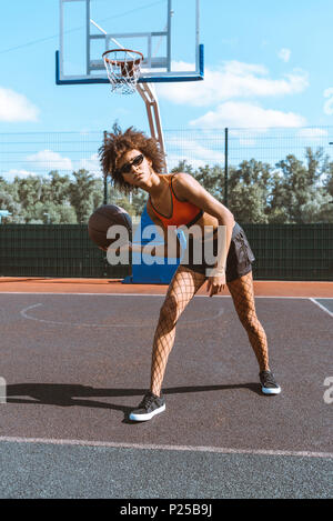 Joven Mujer afroamericana en vestimenta deportiva y tacones altos de pie  con un pie en las escaleras de madera y sosteniendo una pelota de  baloncesto Fotografía de stock - Alamy