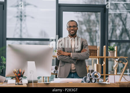 El arquitecto americano africano sonriente con los brazos cruzados en el lugar de trabajo permanente con el ordenador y planos
