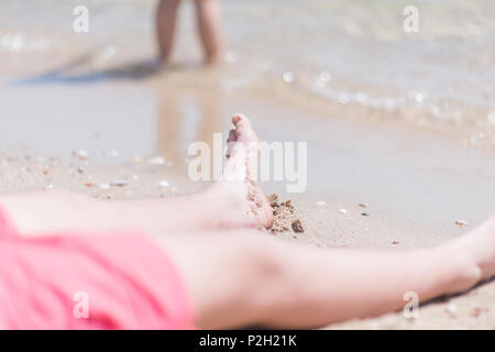 Chico vistiendo el traje de baño rojo descansando sus pies en la arena de la playa en frente del agua