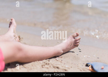 Chico vistiendo el traje de baño rojo descansando sus pies en la arena de la playa en frente del agua