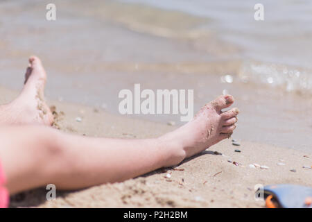 Chico vistiendo el traje de baño rojo descansando sus pies en la arena de la playa en frente del agua