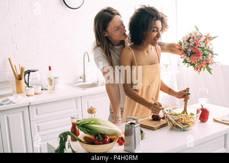 Joven regalar ramo de flores para la novia afroamericanos mientras ella cocina en la cocina