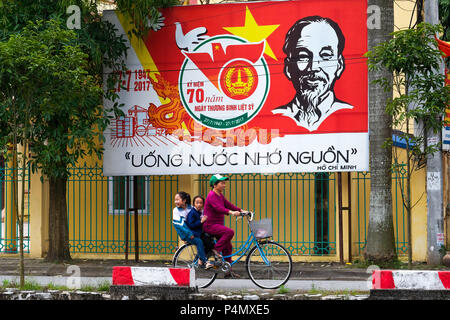 Mujer Bicicleta bajo un póster de propaganda del partido comunista de Vietnam en la provincia de Nam Dinh, Vietnam - Propagandaplakat der Kommunistischen Partei der Dinh-Provinz Vietnams en Nam, Vietnam Foto de stock