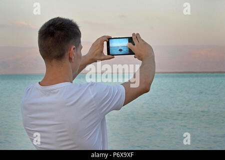 Guy Turista en una camiseta blanca toma fotografías utilizando un smartphone de piedra en el mar al atardecer. El Mar Muerto, Israel. Paisaje de montaña de ensueño de Jordania