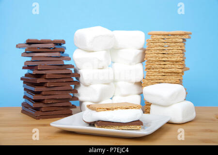 S caseros'more sobre una placa cuadrada con pilas de galletas graham, chocolate y malvaviscos sobre una mesa de madera con fondo de color azul claro. Foto de stock