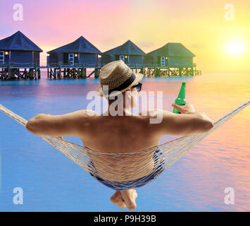 Joven con una botella de cerveza, tumbado en una hamaca en un agua cabañas resort en una isla de Maldivas Foto de stock