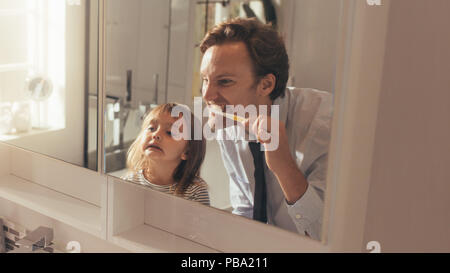 Hombre cepillar sus dientes mirando en el espejo de pie en el baño, mientras que su hija mira. El hombre enseñar a su hija cómo cepillarse los dientes.