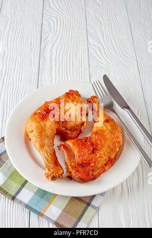 Delicioso pollo frito jugosas piernas con costra de piel dorada sirve sobre una placa blanca con tenedor y cuchillo sobre una tabla de madera,vista vertical desde arriba