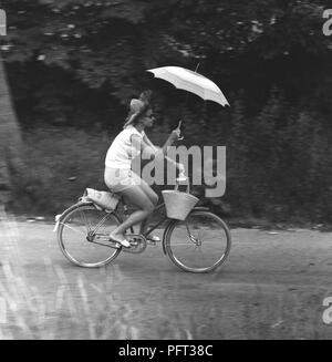 Lluvia en el decenio de 1950. Una mujer joven monta una bicicleta sosteniendo un paraguas para evitar mojarse con la lluvia. Suecia, julio de 1959 Foto de stock