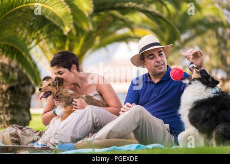 Bonita pareja caucásicos de mediana edad sentados en el césped con palmeras en el fondo mientras jugando con dos perros divertido y hermoso y una bola roja. Disfrute de una