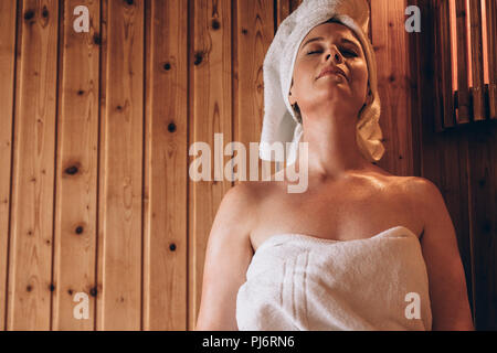 Mujer sentada en una toalla de baño con los ojos cerrados. Mujer relajante en un recinto de madera durante el baño. Foto de stock